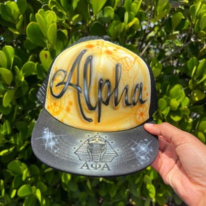 May include: Black and yellow trucker hat with the word "Alpha" in black script. The hat features a black brim with a design of an elephant and pyramid. The background is a lush green.