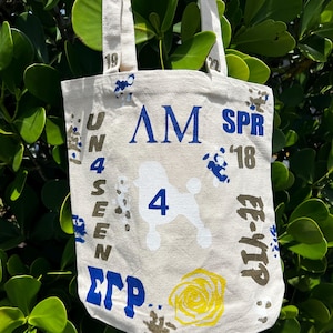 May include: A beige canvas tote bag with a poodle illustration and the text "AM SPR '18". The bag also features the text "UNSEEN" and "ΣΓΡ" in blue and brown. A yellow rose graphic is also visible.