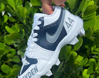 May include: White and navy blue athletic cleat with the name "Zayden" in gray lettering. The number "44" is printed on the side. A silver swoosh logo is visible. The cleat is held against a background of green leaves.
