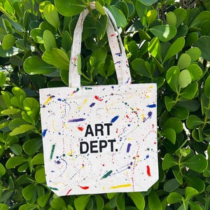 May include: A white canvas tote bag with the words "ART DEPT." in black block letters. The bag is decorated with colorful paint splatters in red, blue, yellow, and green. The bag has white handles.