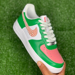 May include: Green and pink custom Nike Air Force 1 sneakers with a white swoosh decorated with pearl-like embellishments. The laces are white and the tongue tag reads "AF1".