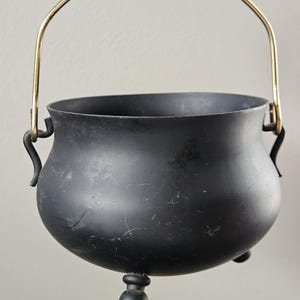 May include: A matte black metal cauldron with a rounded body, three small spherical feet, and a curved gold-toned metal handle attached to two side loops. The surface of the black vessel shows minor signs of wear and light surface scratches.