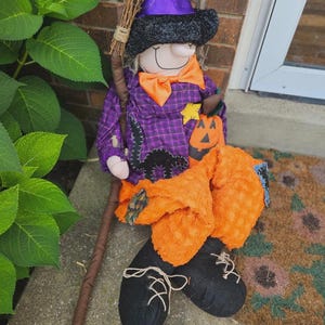 May include: A fabric witch doll wearing a black glittery hat with a purple band, a purple plaid shirt with a black cat applique, an orange bow tie, and orange textured pants. The doll holds a broom and has black shoes with white laces.