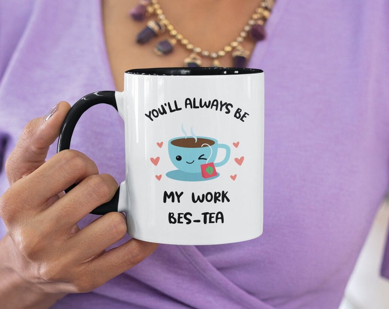 May include: White ceramic mug with a black rim and handle. The mug has a cartoon illustration of a teacup with a smiling face and the text "You'll always be my work bes-tea".