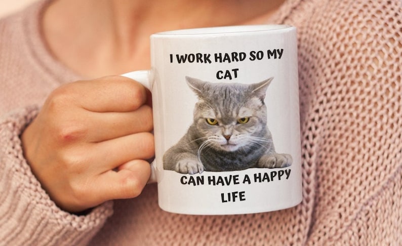 May include: White ceramic mug featuring a grey cat image and the text "I work hard so my cat can have a happy life".