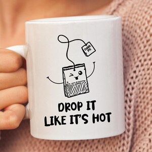 May include: White ceramic mug with a black illustration of a tea bag with a face and the text "Dunk Me" on a tag. The text "Drop it like it's hot" is printed below the tea bag.