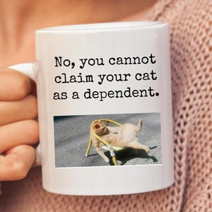 Op de afbeelding: Witte keramische koffiemok met zwarte tekst die "Nee, je kunt je kat niet als een persoon ten laste claimen." luidt en een afbeelding van een kat die in een gele stoel ligt.