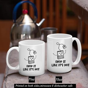 May include: Two white ceramic mugs with black text and illustrations of a tea bag with a face. The smaller mug is 11 ounces and the larger mug is 15 ounces. Both mugs say "Drop it like it's hot".