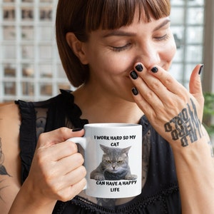 May include: White ceramic mug with a black handle. The mug features a grey cat with a grumpy expression. The text on the mug reads "I work hard so my cat can have a happy life."