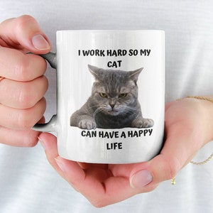 May include: White ceramic mug featuring a black and white image of a cat with the text "I work hard so my cat can have a happy life."