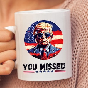 May include: White ceramic coffee mug with a graphic of a man wearing sunglasses and a suit in front of an American flag. The text "You Missed" is printed below the graphic.