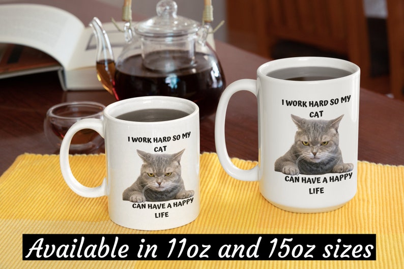 May include: Two white ceramic mugs with black text and a picture of a cat. The text on the mugs reads "I work hard so my cat can have a happy life". The mugs are available in 11oz and 15oz sizes.