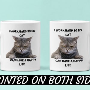 May include: White ceramic coffee mug featuring a grey cat image and the text "I work hard so my cat can have a happy life". The text is printed on both sides of the mug.