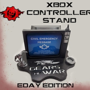Puede incluir: Soporte negro para mando de Xbox con el texto "XBOX CONTROLLER STAND". El soporte presenta una pantalla con el texto "CIVIL EMERGENCY MESSAGE" y el logotipo de Gears of War. La base dice "GEARS OF WAR" y "EDAY EDITION".