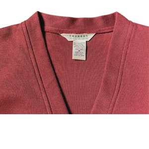 May include: Close-up of a brick-red cardigan with a V-neck. The tag reads "EXPRESS TRICOT" and includes fabric details and care instructions. The garment is size M.