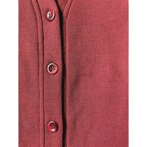 May include: Close-up of a burgundy-colored cardigan with three visible buttons. The buttons are a matching burgundy color and are fastened. The fabric has a ribbed texture.