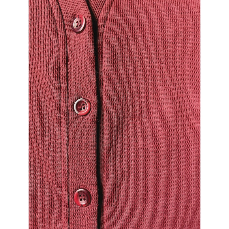 May include: Close-up of a burgundy-colored cardigan with three visible buttons. The buttons are a matching burgundy color and are fastened. The fabric has a ribbed texture.