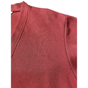 May include: Close-up of a maroon-colored V-neck shirt. The fabric has a ribbed texture, and the stitching is visible along the neckline and shoulder seam. The shirt appears to be made of a soft, comfortable material.
