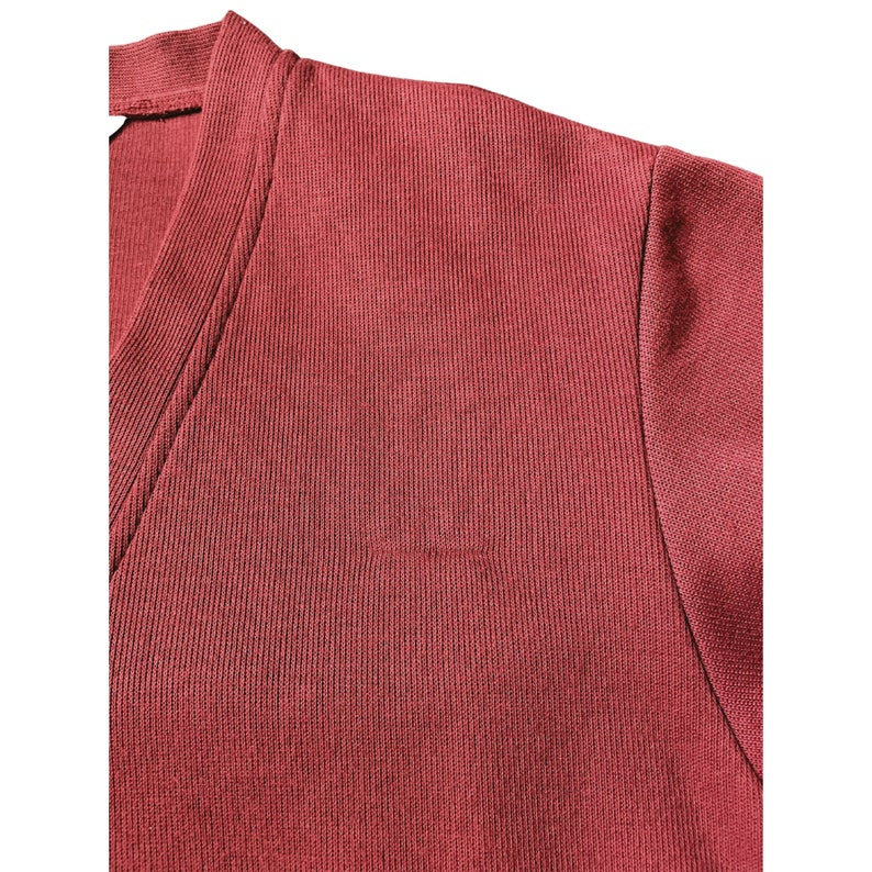 May include: Close-up of a maroon-colored V-neck shirt. The fabric has a ribbed texture, and the stitching is visible along the neckline and shoulder seam. The shirt appears to be made of a soft, comfortable material.