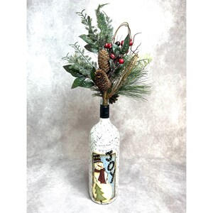 May include: A decorative wine bottle with a snowman design and the word "JOY". The bottle is topped with a festive arrangement of pine, berries, and pinecones, creating a holiday-themed centerpiece.