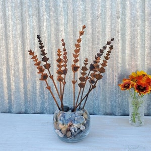 May include: A metal sculpture of a bouquet of lavender flowers in a clear glass vase filled with small stones. The sculpture is rusty brown and has a rustic look.