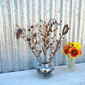May include: A clear glass bowl filled with blue and gray stones holds metal floral sculptures. A small glass vase with orange and yellow flowers sits to the right. The background is a corrugated metal sheet.