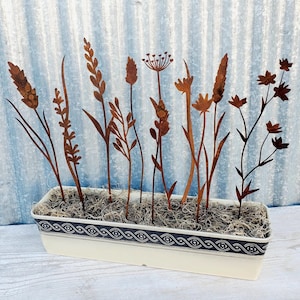 May include: A metal planter with a white base and a black and white patterned rim. The planter is filled with brown moss and contains a variety of metal flower silhouettes in rusty brown.