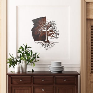 May include: A metal wall art sculpture of a tree with roots and branches, half of the tree is a rusty brown color and the other half is white. The sculpture is hanging on a white wall above a wooden dresser with two drawers.
