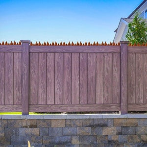 May include: A wooden fence with a brown finish, featuring vertical planks and decorative pointed tops. The fence is set atop a stone retaining wall, with a clear blue sky in the background. A house is visible in the background.
