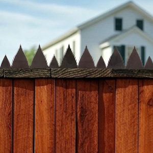 May include: A wooden fence with a row of metal spikes on top. The fence is brown and the spikes are black. The fence is in front of a white house with a blue sky in the background.