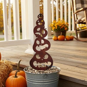 May include: A rustic metal pumpkin stack decoration in a galvanized bucket filled with stones. The stacked pumpkins have cut-out faces and are displayed on a wooden porch with pumpkins and hay bales, creating a fall harvest theme.