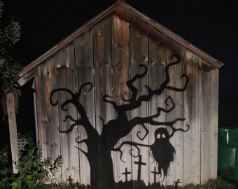 May include: A Halloween metal yard decoration depicting a spooky tree and ghost silhouette. The tree and ghost are mounted on a wooden shed. The yard art is placed in front of a grassy area.