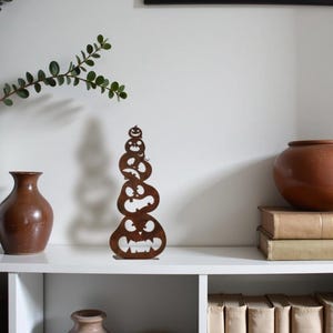 May include: A decorative metal sculpture of stacked jack-o'-lanterns in a warm brown hue. The sculpture is placed on a white shelf, alongside a brown vase, a white vase with greenery, and a framed picture.