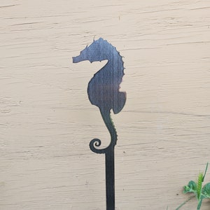 May include: A dark seahorse garden stake. The seahorse is cut from a dark material and has a long stem with a curled design at the bottom. The seahorse is facing left and is set against a light-colored background.