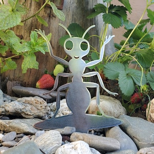 May include: A metal ant garden sculpture with outstretched arms, set among rocks and plants. The ant has a gradient color scheme, transitioning from silver to dark gray. Strawberries and greenery surround the sculpture, creating a natural garden scene.