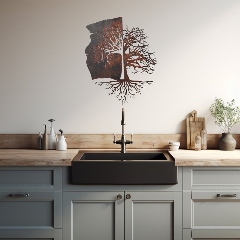 May include: A rustic metal tree wall art with a split trunk and intricate roots. The tree is mounted on a white wall above a black farmhouse sink in a kitchen.
