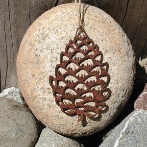 May include: A rustic, brown metal pinecone ornament with a mountain silhouette design, hanging by a natural twine string. The ornament is displayed on a large, smooth, light-colored stone, suggesting a nature-inspired theme.