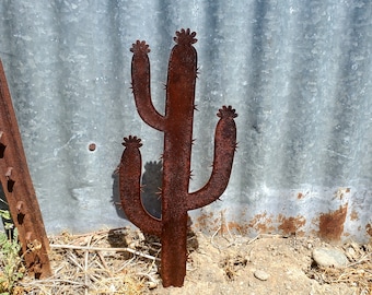 May include: A rusty metal cactus sculpture with three arms and spiky details. The desert-themed art piece stands in front of a corrugated metal background, adding a rustic touch to any outdoor space. The cactus is approximately 30 inches tall.