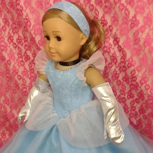 Puede incluir: Una muñeca vestida con un vestido azul claro, una diadema a juego y guantes plateados. El vestido tiene una falda en capas y un corpiño detallado. La muñeca tiene el pelo rubio y está colocada sobre un fondo de encaje rosa.