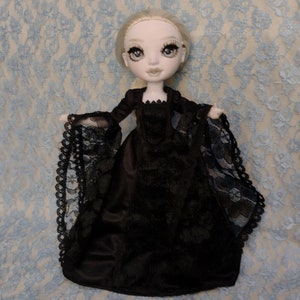 Könnte beinhalten: Handgefertigte Puppe mit heller Haut, blonden Haaren und großen Augen, bekleidet mit einem schwarzen Kleid mit Spitzenärmeln und -besatz. Die Puppe ist vor einem hellblauen Spitzenstoffhintergrund platziert.