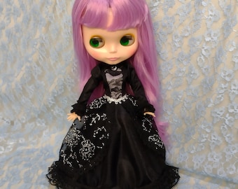 May include: A doll with long purple hair and large green eyes, wearing a black dress with silver accents and lace detailing. The doll is posed against a light blue, floral-patterned fabric.