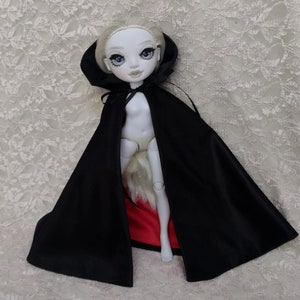 Könnte beinhalten: Eine Puppe mit heller Haut, blonden Haaren und blauen Augen, die einen schwarzen Umhang mit rotem Futter trägt. Die Puppe ist vor einem strukturierten, cremefarbenen Hintergrund positioniert, was eine gotische oder Fantasy-Ästhetik erzeugt.