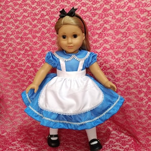 Puede incluir: Muñeca vestida con un vestido azul con un delantal blanco, que recuerda a Alicia en el país de las maravillas. El vestido tiene ribetes de encaje blanco y mangas cortas. La muñeca tiene cabello rubio, una diadema negra y zapatos negros.