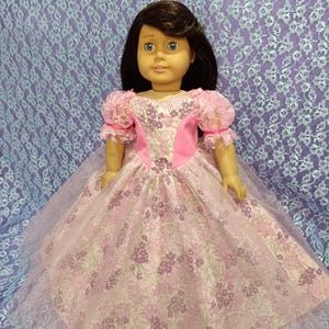 Puede incluir: Una muñeca con un vestido floral rosa con una capa de tul. El vestido tiene un corpiño y mangas rosas con ribete de encaje. La muñeca tiene cabello castaño y ojos azules. El fondo es un patrón de encaje lila claro.