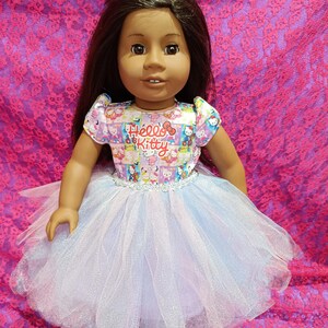 Puede incluir: Una muñeca con cabello castaño oscuro que lleva un vestido de Hello Kitty con una falda de tul azul claro. El vestido tiene un estampado blanco, rosa y azul con las palabras "Hello Kitty". La muñeca está sobre un fondo de encaje rosa.