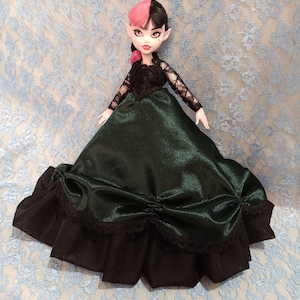 May include: A doll with pink and black hair, dressed in a black lace top and a dark green satin dress with black lace trim. The doll has pale skin and is set against a light blue lace background.