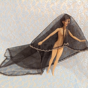 Puede incluir: Una muñeca de moda con cabello oscuro y un velo negro. El velo está adornado con encaje dorado y cubre el cuerpo de la muñeca. La muñeca está desnuda, con articulaciones, y posa sobre un fondo floral azul claro y blanco.