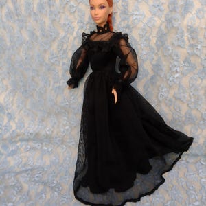 May include: A doll in a long black dress with sheer sleeves and a high neckline. The dress features a fitted bodice and a flowing skirt. The doll has red hair and is set against a light blue lace backdrop.