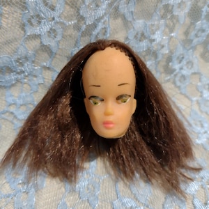 May include: A vintage doll's head with brown hair and painted facial features. The doll has green eyes, thin eyebrows, and a pink mouth. The head is made of a light-coloured material and is surrounded by long, brown hair.