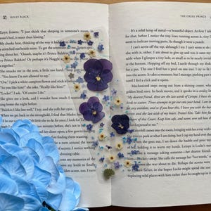 Könnte beinhalten: Ein transparentes, rechteckiges Lesezeichen mit eingearbeiteten gepressten violetten Stiefmütterchen sowie kleinen blauen und weißen Blüten. Das Lesezeichen liegt auf einer offenen Buchseite, im Vordergrund ist eine große hellblaue Hortensie zu sehen.